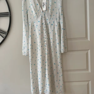 Vit långklänning med blå blommor - Superfin långklänning i vit satinliknande kvalitet med små blå blommor från Adoore. Klänningen har långa ärmar, v-ringning och knappar framtill. Perfekt för dig som gillar romantisk och feminin stil med en touch av vintage. Helt ny som aldrig hann bli använd och nu inte passar mig tyvärr. 