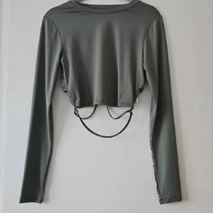 Grågrön croppad långärmad topp FB Sister - Snygg grågrön croppad topp från FB Sister med långa ärmar och coola snörningsdetaljer i midjan. Toppens material är mjukt och stretchigt, perfekt för en trendig look. Passar dig som gillar stilrena plagg med en twist.