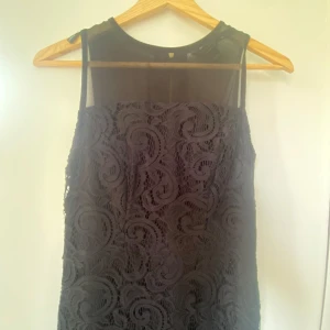 Svart spetsklänning med dragkedja - Svart långklänning  med snygg spets och transparent parti upptill. Klänningen är ärmlös och har en guldig dragkedja i ryggen som ger en cool detalj. Perfekt för dig som gillar stilrena och eleganta plagg med en twist.