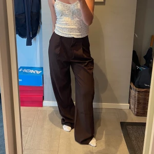 Bruna vida kostymbyxor - Snygga bruna kostymbyxor med hög midja och vida ben. Byxorna har pressveck framtill, sidfickor och fuskfickor bak. Perfekta för en trendig och avslappnad look. Materialet känns mjukt och följsamt. Over sized fit. Från NAKD. Använd kanske 2 ggr.