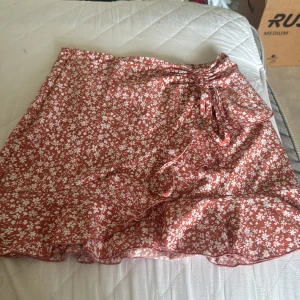 Blommig omlottkjol med volang - Supersöt kort omlottkjol i rödbrun nyans med vita blommor. Kjolen har volangkant och knyts i midjan för en snygg passform. Perfekt för varma dagar när du vill känna dig både chill och stylish.