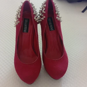 Röda pumps med nitar och strass - Riktigt snygga röda pumps från Newtrend med hög klack och platåsula. Baksidan och klacken är täckta av silvriga nitar och strass för en edgy look. Skorna har rund tå och är klädda i ett mockaliknande material.