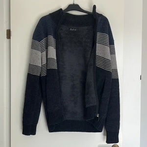 Mörkblå randig kofta med dragkedja - Mysig mörkblå kofta med grå och vita ränder. Koftan har hög krage, hel dragkedja och två fickor framtill. Insidan är fodrad med mjuk fleece för extra värme. Perfekt för kyliga dagar och snygg till jeans.