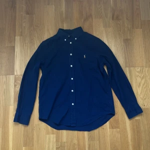 Mörkblå skjorta från Ralph Lauren, storlek 14-16 - Klassisk mörkblå skjorta från Ralph Lauren med button-down krage och vita knappar. Skjortan har lång ärm och den ikoniska broderade loggan på bröstet. Perfekt för en clean och stilren look.