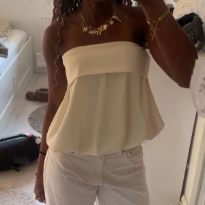 Ljusbeige bandeau tubtopp - Säljer en superfin ljusbeige bandeau tubtopp med luftig och flowy passform. Toppen är axelbandslös och har en snygg volangliknande kant upptill. Perfekt för varma dagar och enkel att styla med jeans eller kjol.