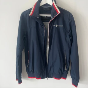 Marinblå vindjacka från Sail Racing - Snygg marinblå vindjacka från Sail Racing med röda och vita detaljer på krage och muddar. Jackan har dragkedja framtill, två fickor med dragkedja och grått foder. Broderad logga på bröstet och patch på ärmen ger en sportig vibe.