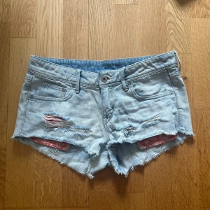 Ljusblåa lågmidu jeansshorts  - Snygga ljusblå jeansshorts från gamla Hm med slitna detaljer, fransig kant och röda bandana-tygdetaljer i hålen framtill. Klassisk femficksmodell och låg midja