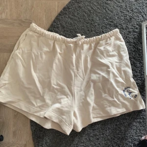 Beige mjuka shorts med snörning - Superbekväma beige shorts med elastisk midja och snörning. Tillverkade i mjukt bomullstyg och har en liten blå logga på ena benet. Perfekta för chill dagar eller träning. Lätta att matcha med en hoodie eller t-shirt.