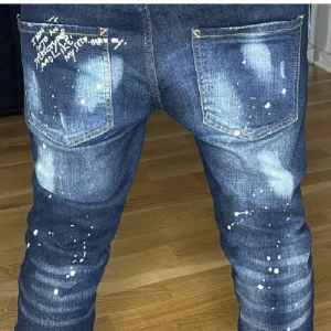 Dsquared2 blå jeans med slitningar - Säljer ett par blå jeans från Dsquared2 med coola slitningar, färgstänk och textdetaljer på bakfickan. Jeansen har en smal passform och passar nån som är runt 180