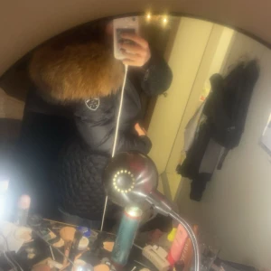 Svart dunjacka med stor pälsluva - Säljer en svart dunjacka med stor fluffig pälsluva och quiltad design. Jackan har en badge på ärmen och är perfekt för kalla dagar. Snygg och varm med lång ärm och markerad midja. Passar dig som vill ha en stilren vinterlook.