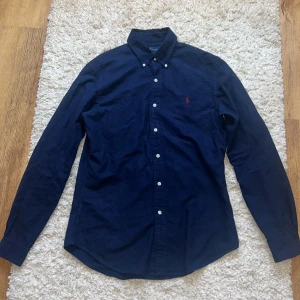 Mörkblå skjorta från Ralph Lauren M - Säljer en mörkblå slim fit skjorta från Ralph Lauren i bomull. Klassisk button-down krage, vita knappar och rött broderat Polo-logo på bröstet. Långärmad och stilren modell som passar perfekt till jeans eller chinos.