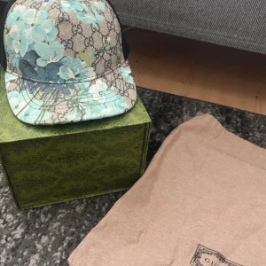 Gucci keps med blå blommor - Säljer en exklusiv Gucci keps i storlek M med ikoniskt GG-mönster och stora blå blommor framtill. Kepsen har böjd skärm, mesh på baksidan och klassisk logotypetikett på insidan. Perfekt för dig som vill sticka ut med en lyxig accessoar. Fint skick, osäker på äkthet men fick online kvitto.
