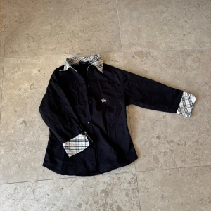 Svart Burberry skjorta med rutiga detaljer - Svart skjorta från Burberry med klassiskt rutigt mönster på krage och ärmslut. Skjortan har långa ärmar, knappar framtill och ikonisk broderad ryttare på bröstet. Tillverkad i bomull och har en normal passform. Perfekt för dig som gillar stilrena och tidlösa plagg. Stl M i !barn storlekar! ÄKTA