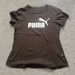 Svart Puma t-shirt med logga - Svart t-shirt från Puma med stor vit logga och text på bröstet. Klassisk rund halsringning och korta ärmar. Tillverkad i mjuk bomull som känns skön mot huden. Perfekt för dig som gillar sportiga och stilrena plagg.