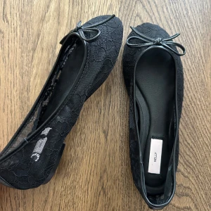 Nya Svarta spetsballerinor från Nelly - Snygga svarta ballerinaskor från Nelly med spetsdetaljer och liten rosett framtill. Skorna har rund tå och platt sula, vilket gör dem både stilrena och bekväma. Perfekta för dig som gillar en feminin och trendig look. Oanvända