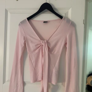 Ljusrosa omlottopp från Gina Tricot - Supersöt ljusrosa omlottopp från Gina Tricot med lång ärm och knyt framtill. Toppens v-ringning och mjuka bomullsmaterial gör den både bekväm och trendig. Perfekt att styla med jeans eller kjol för en fräsch look.