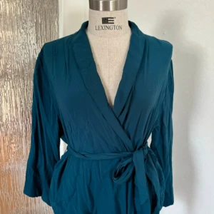 Omlottblus The Shirt Factory - Snygg mörkblå omlottblus från The Shirt Factory i storlek 38. Blusen har trekvartsärm och knyts i midjan med ett bälte för en smickrande siluett. Perfekt för dig som gillar stilrena och enkla plagg med en twist.
