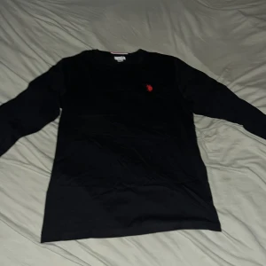 Svart långärmad tröja U.S. Polo Assn. - Svart långärmad tröja från U.S. Polo Assn. med röd broderad logga på bröstet. Klassisk rak passform och rund halsringning. Tillverkad i mjuk bomull som är skön mot huden. Perfekt för dig som gillar stilren och enkel design.