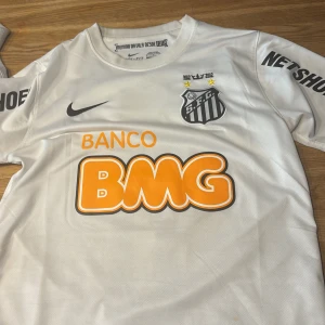Vit Santos Neymar Jr matchtröja Nike S - Santos FC fotbollströja i vitt med Neymar Jr och nummer 11 på ryggen. Tröjan är tillverkad i Dri-Fit-material och har korta ärmar. Nike-logga på bröstet, klubbmärke och flera sponsortryck i orange och svart. Perfekt för dig som älskar brasiliansk fotboll.