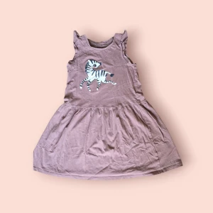 Ljusbrun klänning med zebra-tryck - Supersöt ljusbrun kortklänning i bomull med ett stort zebramotiv framtill. Klänningen är ärmlös med små volangdetaljer vid axlarna och har en utställd kjol. Perfekt för dig som gillar djur och vill ha en lekfull stil.