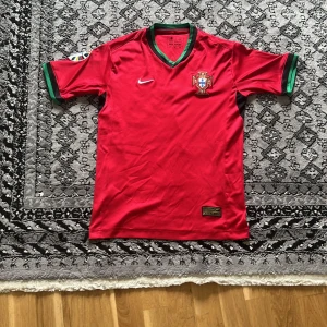 Portugal Ronaldo 7 Nike fotbollströja - Portugal landslagströja med Ronaldo 7 på ryggen. Röd med gröna detaljer vid ärmslut och krage, officiellt Nike-märke och broderat landslagsemblem. Patch på ärmen och guldtryck på ryggen. Tillverkad i lätt och ventilerande material.