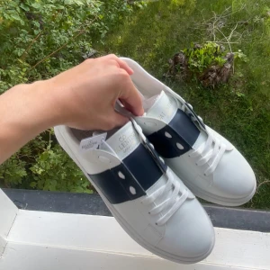  vita sneakers - Snygga vita sneakers med bred marinblå rem över vristen och guldfärgad logga på plösen. Skorna har klassisk snörning, rund tå och är tillverkade i skinn med en chunky, mönstrad sula. Perfekta för dig som vill ha en clean men lyxig look.