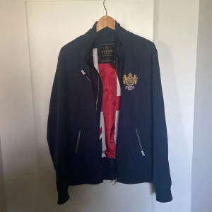 Morris marinblå vindjacka med brodyr - Snygg marinblå vindjacka från Morris med guldbroderad logga och brittisk flagga på bröstet. Jackan har dragkedja, ribbade muddar och två fickor med dragkedja. Insidan är fodrad i rött och vitt för en extra touch. Perfekt för dig som gillar klassisk stil.