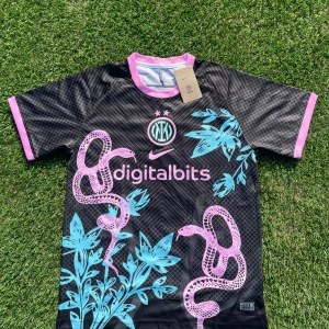 Inter Milan x Nike svart fotbollströja - Inter Milan fotbollströja från Nike i svart med rosa och turkosa detaljer. Tröjan har korta ärmar, rosa muddar och krage, samt unikt mönster med ormar och växter. Digitalbits-logga på bröstet och klubbmärke i turkos och rosa. Tillverkad i lätt och ventilerande polyester.