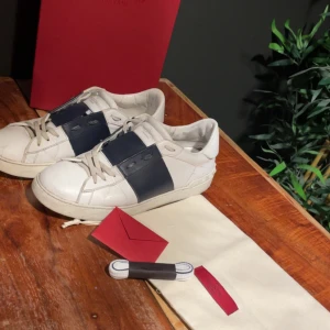 Valentino Open  - Snygga Valentino Garavani Open sneakers i vitt skinn med bred marinblå rem över vristen. Storlek 42 men passar 43. Klassisk rund tå, platt sula och diskret logga på plösen. Skorna har snörning och coola detaljer baktill. Perfekta för dig som gillar exklusiva och stilrena sneakers. Priset är diskuterbart, billigare vid snabba affärer.