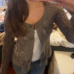 Grå spetskofta med knappar - Supersnygg grå kofta med genombruten spets och stora knappar framtill. Koftan har långärmade ärmar och markerade kanter i tyg. Perfekt att slänga över ett linne eller t-shirt för en chill men ändå uppklädd look. Saknas 1 knapp men man lägger knappt märke till det