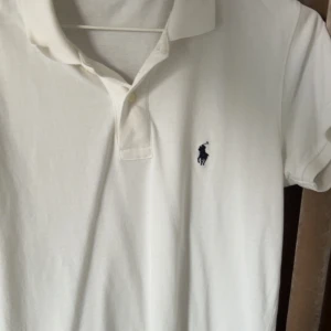 Vit pikétröja från Ralph Lauren - Klassisk vit pikétröja från Ralph Lauren med broderad marinblå logga på bröstet. Tröjan har korta ärmar, krage och knäppning framtill. Perfekt för en clean och stilren look.