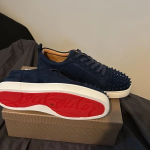 Mörkblå Louboutin sneakers med nitar - Snygga mörkblå sneakers från Louboutin i mocka med ikoniska röda sulor och coola nitar över tån. Skorna har snörning och vit platt sula. Perfekta för dig som vill sticka ut med exklusiv streetstyle.