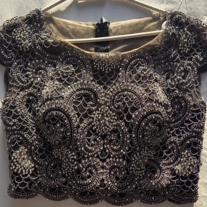 Festklänning 2pieces, Svart kjol med en glittrig topp - Supersnygg svart klänning (kjol + topp) med glittriga pärlor och paljetter i ett detaljerat mönster. Croppad modell med korta ärmar och rund halsringning. Perfekt för att sticka ut på festen! Stängs med dragkedja bak och har transparent mesh i basen.