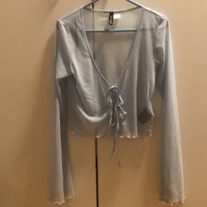 Ljusblå croppad topp från H&M Divided - Supersöt ljusblå croppad topp från H&M Divided med långa ärmar och v-ringning. Toppen har knyt framtill och vågig kant nertill för en extra trendig look. Perfekt att styla med jeans eller kjol för en chill vibe.