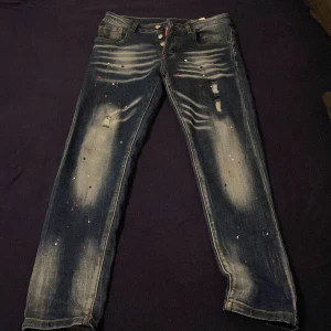 Dsquared2 blå slim jeans med patch - Snygga blå jeans från Dsquared2 med coola slitningar, färgstänk och patchdetaljer bak. Slim fit-modell med flera knappar i gylfen och tydliga Dsquared2-loggor. Jeansen har en unik look med stora röda och gula patches på bakfickan.