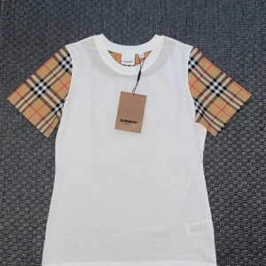  Burberry t-shirt - Snygg vit t-shirt från Burberry med klassiska beige-rutiga ärmar. STORLEK M MEN PASSAR S MER! Perfekt för dig som vill ha en stilren look med en touch av lyx. ALDRIG ANVÄND.