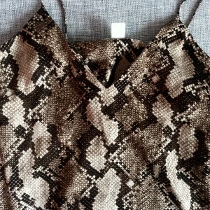 Brun ormmönstrad linnetopp H&M - Säljer en snygg linnetopp från H&M i storlek 36 med ormmönster i bruna och beige toner. Tunt tyg med smala axelband och v-ringning. Perfekt för dig som vill sticka ut med ett djurmönster och gillar trendiga detaljer.