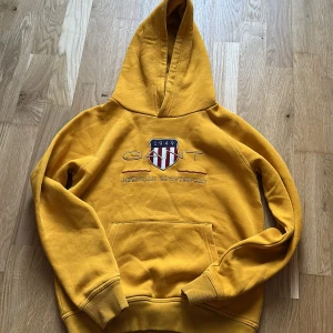Gul hoodie från Gant med brodyr - Gul hoodie från Gant med broderad logga och sköld på bröstet. Klassisk känguruficka framtill, ribbade muddar och huva. Mjuk och skön bomullskvalitet, perfekt för chill dagar eller när du vill sticka ut lite extra.