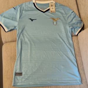 Ljusblå Lazio fotbollströja  - Snygg ljusblå Lazio fotbollströja från med klubbmärke på bröstet och diskreta mönster i tyget. Tröjan har korta ärmar, svart och vit kant vid hals och ärmslut samt klassisk sportig passform. Perfekt för dig som älskar italiensk fotboll.