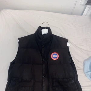 Svart dunväst från Canada Goose M - Svart dunväst från Canada Goose med hög krage, stora framfickor och klassisk logga på bröstet. Västen har både dragkedja och knappar framtill. Perfekt för lager-på-lager och riktigt snygg till streetwear eller outdoor-stil.