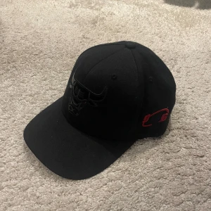 Svart Chicago Bulls keps Mitchell & Ness - Svart keps från Mitchell & Ness med Chicago Bulls-logga broderad framtill i svart och röd brodyrdetalj på sidan och bak. Klassisk snapback-modell med böjd skärm och justerbar passform. Perfekt för dig som gillar basket och streetstyle.