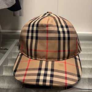 Burberry keps - Snygg keps från Burberry med klassiskt rutigt mönster i beige, svart, vitt och röda linjer. Kepsen har böjd skärm och justerbar rem bak med Burberry London England-logga. Tillverkad i bomull och polyester, perfekt för att lyfta din outfit.