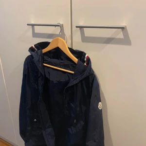 Mörkblå vindjacka från Moncler - Snygg mörkblå vindjacka från Moncler med huva och dragkedja. Jackan har Moncler-logga på ärmen och coola rödvita detaljer på huvan. Tillverkad i lätt polyester som passar perfekt när det blåser ute. Enkel och stilren design som funkar till det mesta.