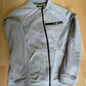 Grå Sail Racing jacka med dragkedja - Snygg grå jacka från Sail Racing med svart dragkedja och logga på bröstet. Jackan har lång ärm och coola reflexdetaljer på ärmarna. Perfekt för dig som gillar stilren och sportig look. Materialet känns mjukt och följsamt.