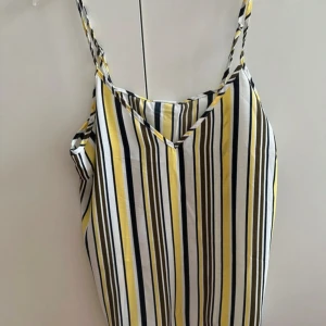 Randig sommar topp med smala axelband - Snygg topp med smala axelband och v-ringning. Toppen har ett randigt mönster i vitt, gult, svart och olivgrönt. Perfekt för varma dagar och ger en fräsch och färgglad look. Lätt och luftig känsla