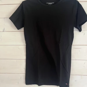 Svart t-shirt från Tommy Hilfiger - En klassisk svart t-shirt från Tommy Hilfiger i mjuk bomull. T-shirten har korta ärmar, rund halsringning och en liten diskret logga nertill. Perfekt basic-plagg med snygg passform och stilren look.