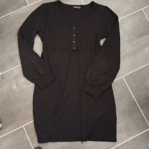 Svart stickad klänning med knappar - Mysig svart stickad klänning från Bonaparte med långa ärmar, ribbad överdel och knappar framtill. Klänningen har snörning i midjan och en rund halsringning. Perfekt för lager-på-lager och chill dagar.