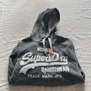 Mörkgrå Superdry hoodie med tryck - Snygg mörkgrå hoodie från Superdry med stort vitt broderat logotyp-tryck och japanska tecken på bröstet. Tröjan har huva med snörning, känguruficka och ribbade muddar. Perfekt för chill dagar eller när du vill ha en casual streetstyle. Vid armen har den en logotyp vilket gör den ännu fetare. 