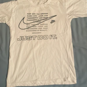 Vit Nike t-shirt med tryck - Vit t-shirt från Nike med klassiskt svart tryck framtill, inklusive den ikoniska swoosh-loggan och texten 'JUST DO IT.' Skön passform och rund halsringning. Perfekt för dig som gillar sportig och clean stil. Det är storlek S