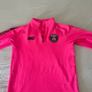 Rosa PSG långärmad träningströja Nike - Snygg långärmad fotbollströja från Nike i stark rosa färg med PSG-logga på bröstet och svart Nike-tryck. Tröjan har halv dragkedja framtill och svarta detaljer längs armarna där det står 'PARIS' och Nike-loggor. Perfekt för träning eller när du vill sticka ut.